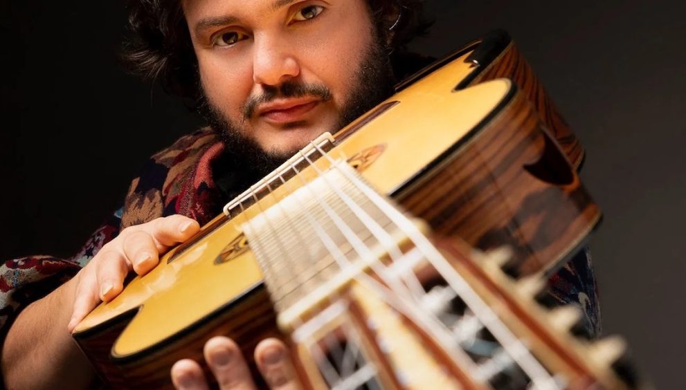 Yamandu Costa holding acoustic guitar, looking down the guitar neck into the camera
