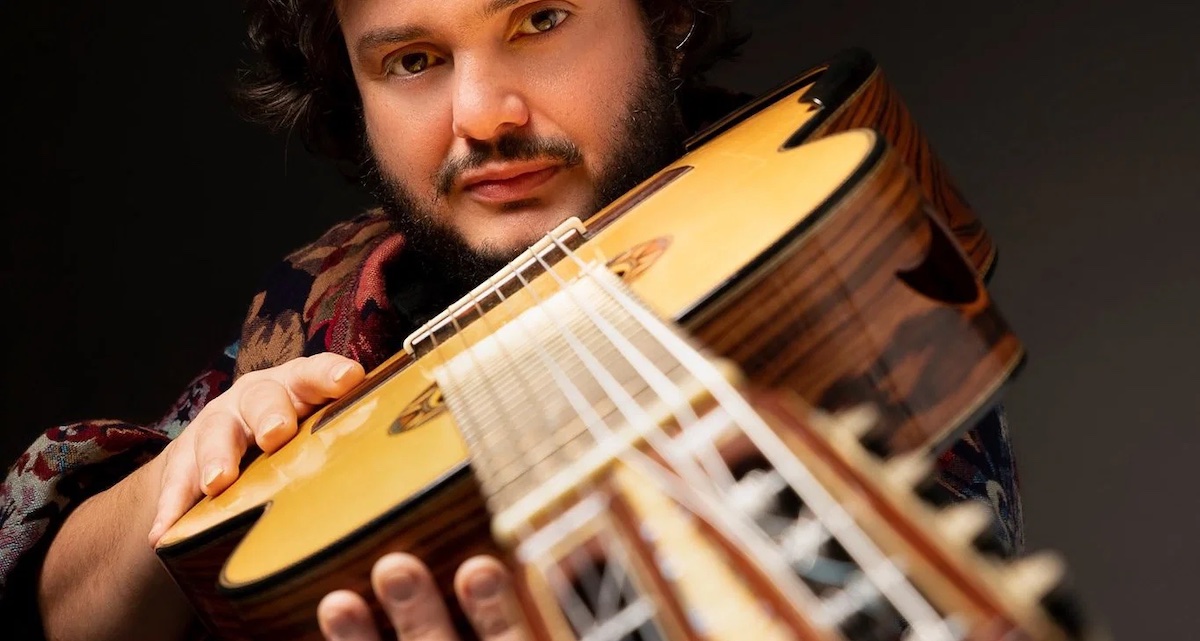 Yamandu Costa holding acoustic guitar, looking down the guitar neck into the camera