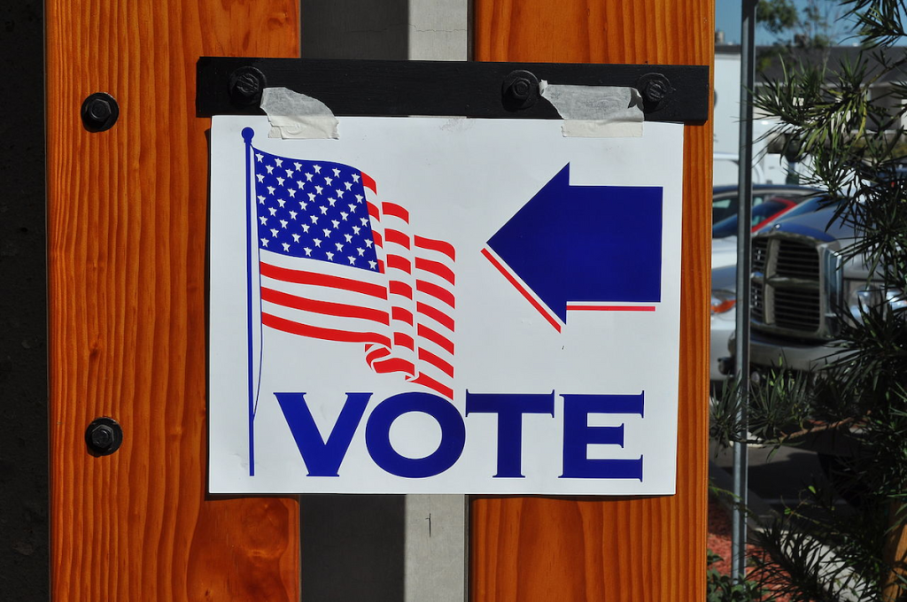 white sign has American (U.S.) flag and the word "vote" and an arrow in blue