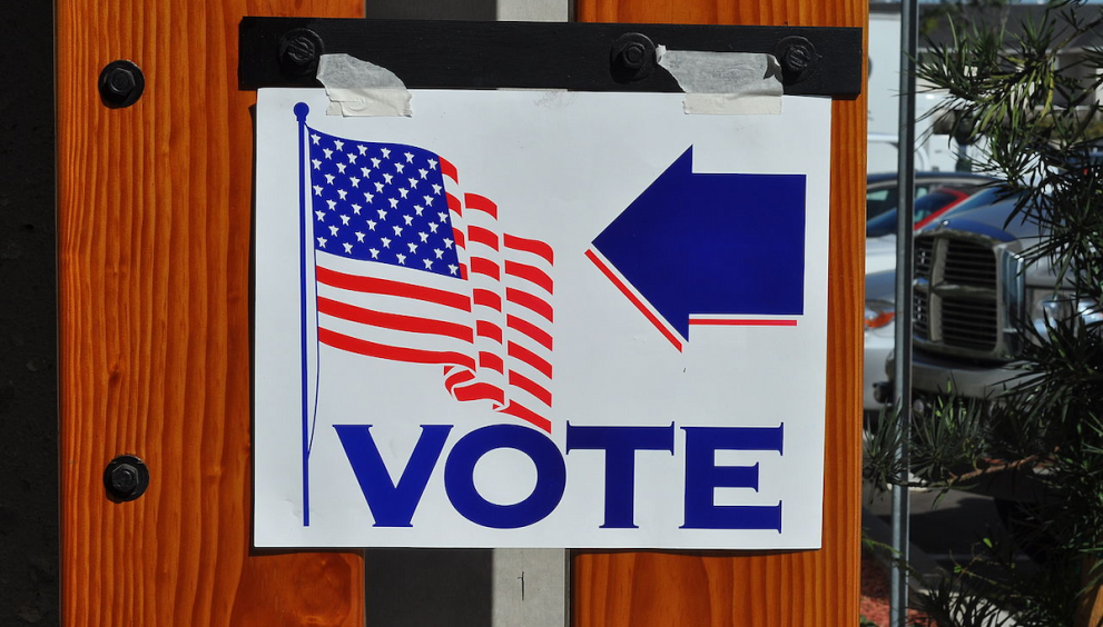 white sign has American (U.S.) flag and the word "vote" and an arrow in blue