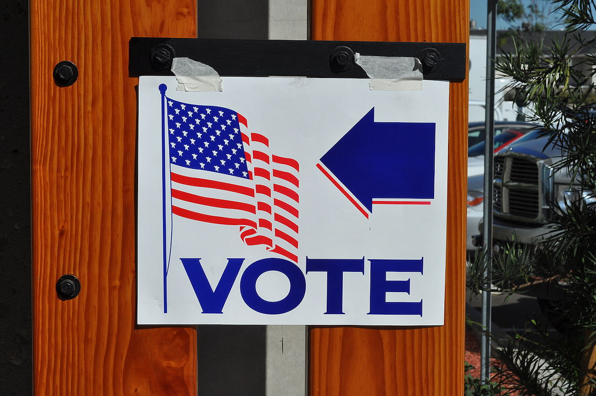 white sign has American (U.S.) flag and the word "vote" and an arrow in blue