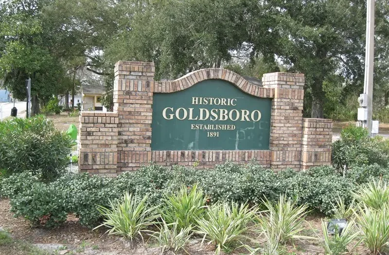 A detailed brick entrance sign marks the community of 'HISTORIC GOLDSBORO ESTABLISHED 1891' in Sanford, Florida. It is set amidst green foliage and mature oak trees.