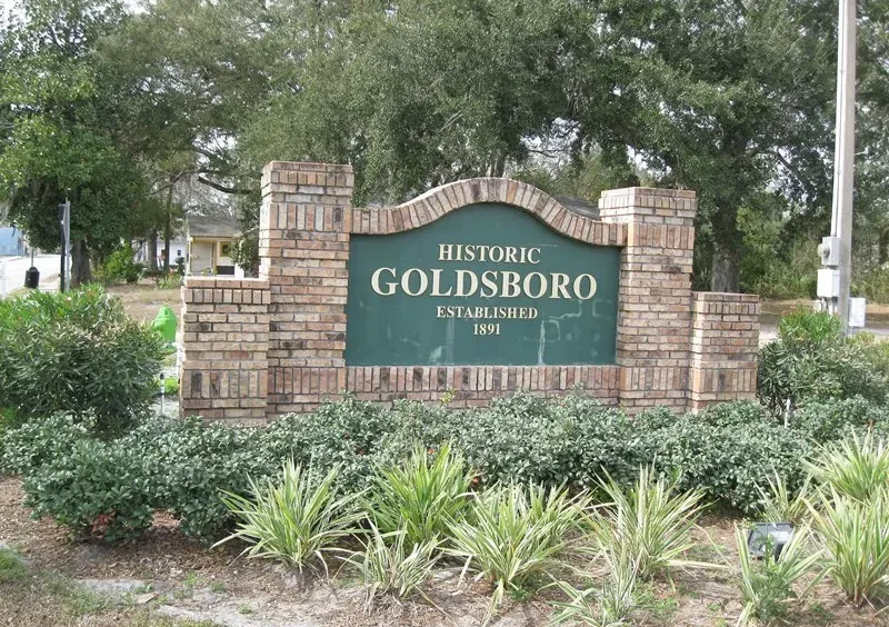 A detailed brick entrance sign marks the community of 'HISTORIC GOLDSBORO ESTABLISHED 1891' in Sanford, Florida. It is set amidst green foliage and mature oak trees.