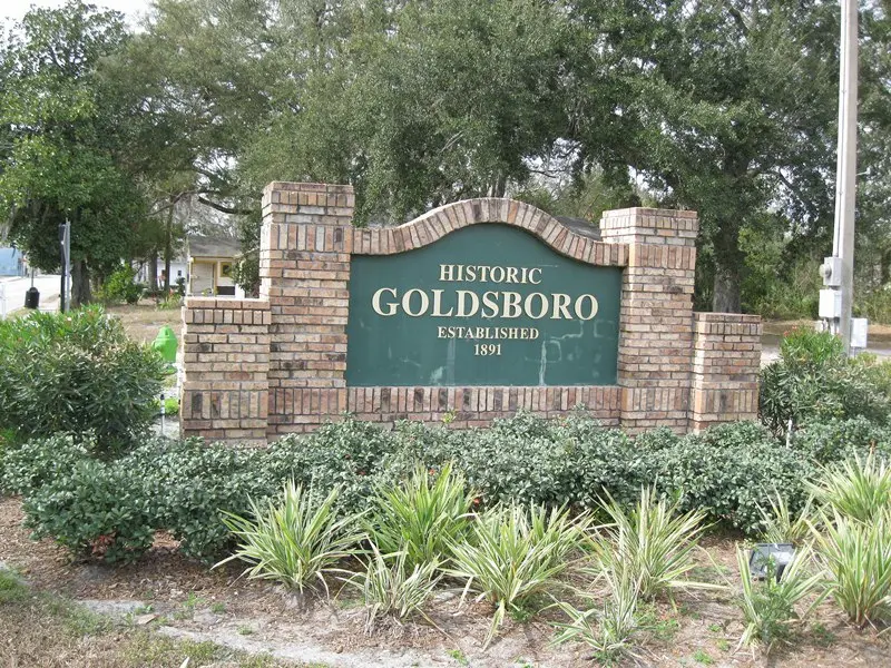 A detailed brick entrance sign marks the community of 'HISTORIC GOLDSBORO ESTABLISHED 1891' in Sanford, Florida. It is set amidst green foliage and mature oak trees.
