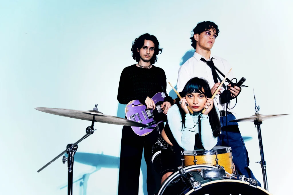 The three members of Orlando indie band Mirror Parts pose with a guitar, drums, and microphone in promotional photo against light blue background.