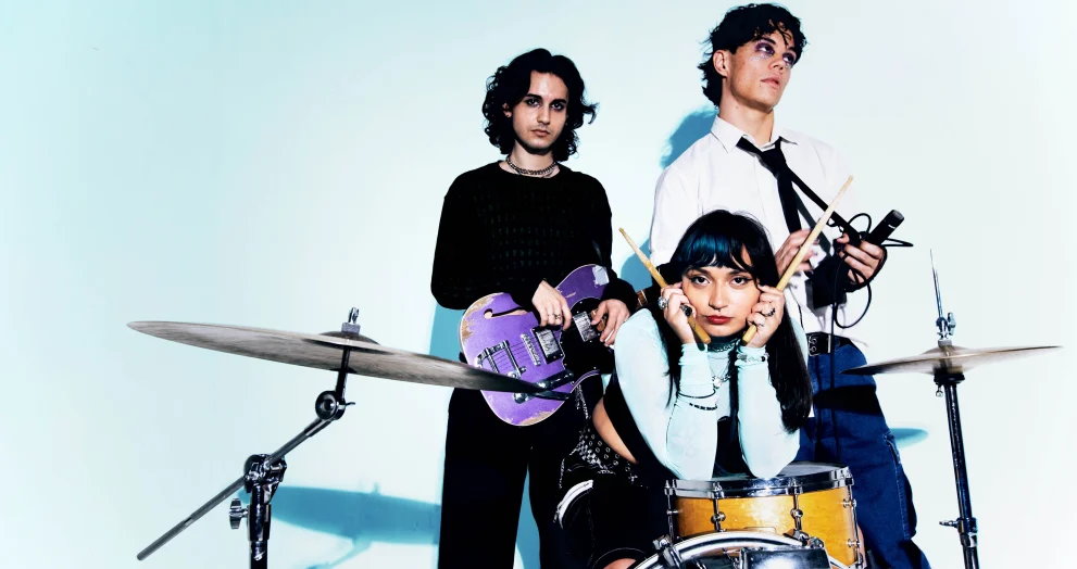 The three members of Orlando indie band Mirror Parts pose with a guitar, drums, and microphone in promotional photo against light blue background.