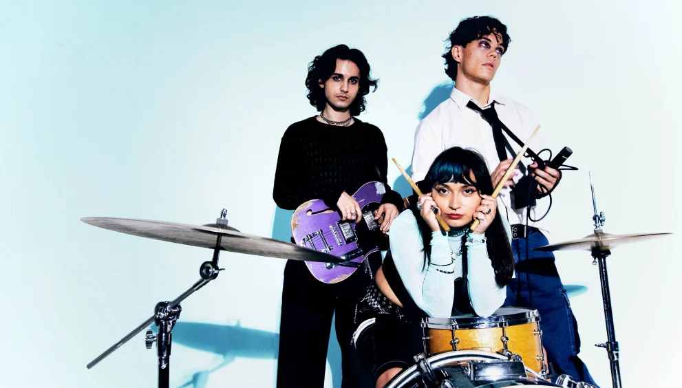 The three members of Orlando indie band Mirror Parts pose with a guitar, drums, and microphone in promotional photo against light blue background.