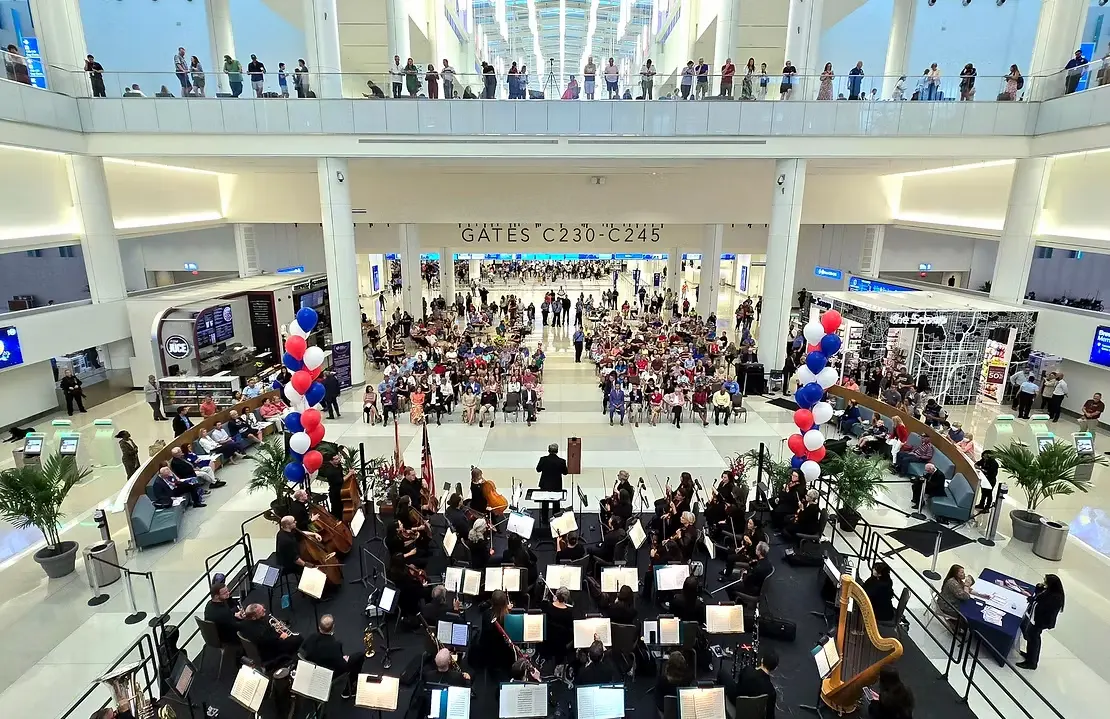 Orchestra performing on a stage inside a large airport terminal, with a conductor and seated audience surrounded by red, white, and blue balloons.