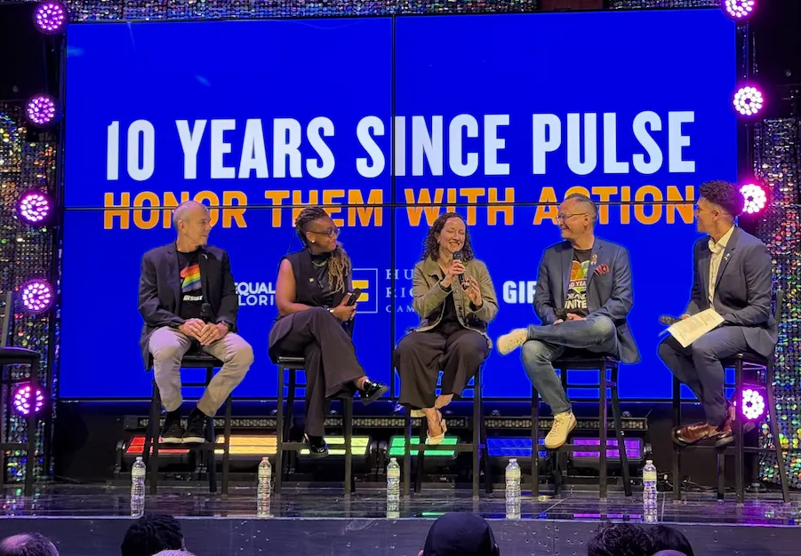 Five people sitting on stools on a stage participating in a panel discussion in front of a blue screen that reads "10 Years Since Pulse: Honor Them With Action."