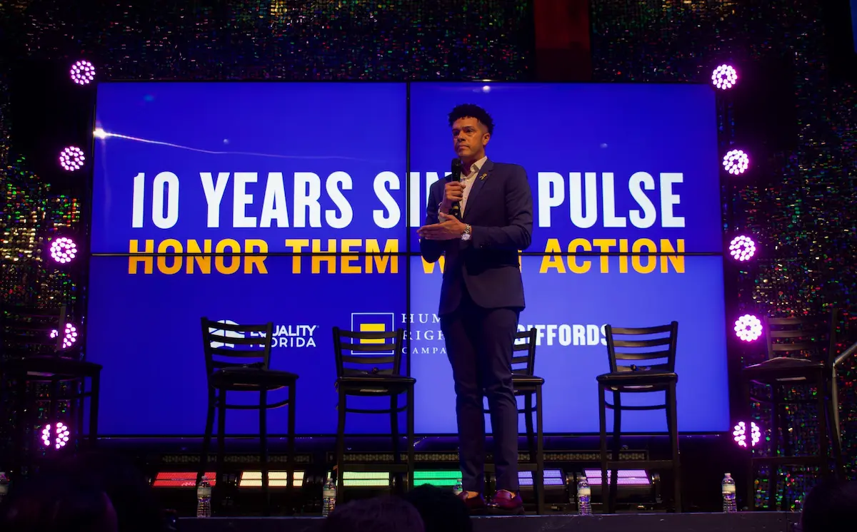 Brandon Wolf, a man wearing a suit, stands on stage with microphone in front of four empty chairs and a backing screen that reads "10 Years Since Pulse: Honor Them With Action"