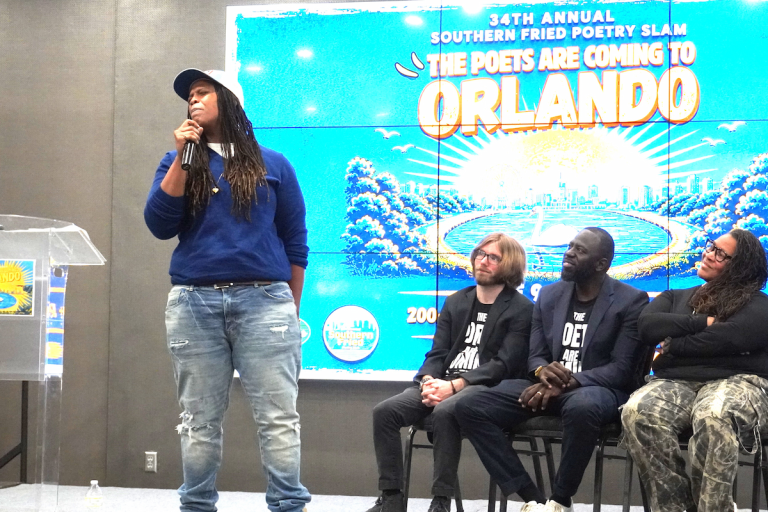 A poet with long black hair wearing blue sweater and jeans on a stage holding a microphone performs dramatically next to a panel of three seated individuals in front of a bright blue digital backdrop reading "34th Annual Southern Fried Poetry Slam: The Poets Are Coming to Orlando."