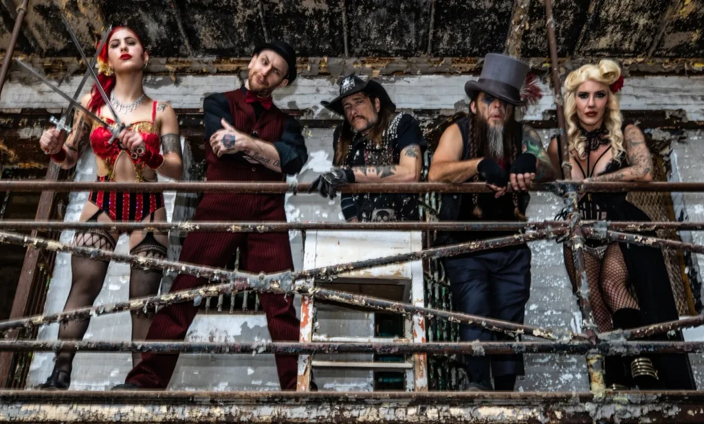 Five heavily tattooed performers in gothic and alternative circus attire (red lingerie with swords, red vest, biker vest, top hat, black corset) standing on a dilapidated metal fire escape against a severely decayed building wall.