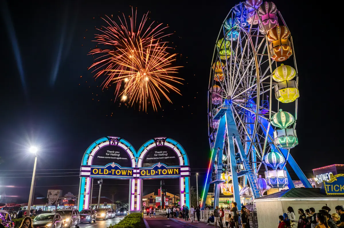 Classic cars lined up for Red White and Horsepower 4th of July event at Old Town Kissimmee. Large colorful ferris wheel stands tall against a dark night sky as am orange firework is deployed.