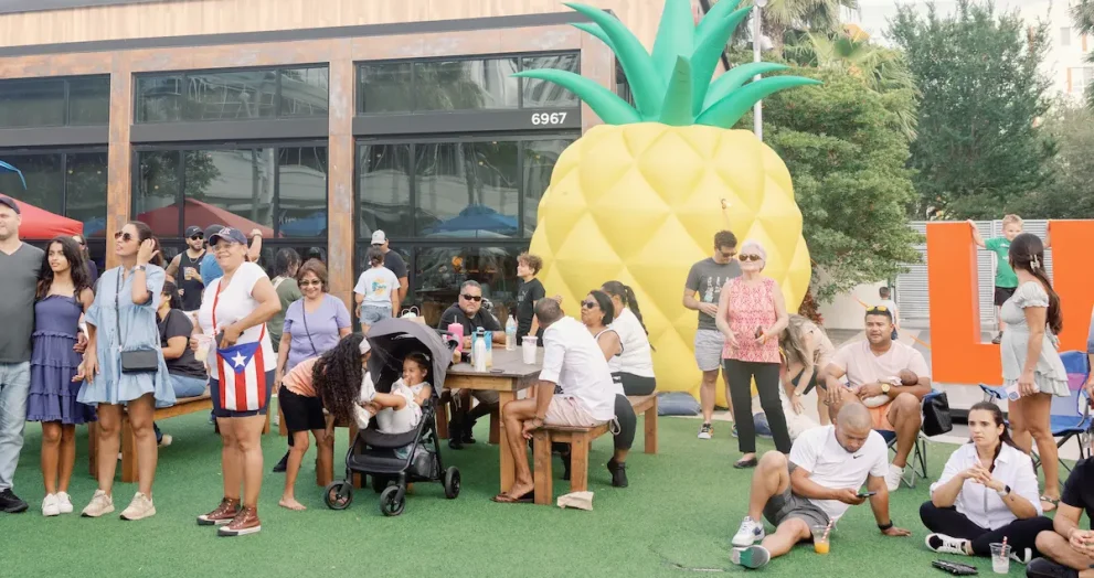 people outdoors sitting and standing around large inflatable pineapple, enjoying entertainment and food