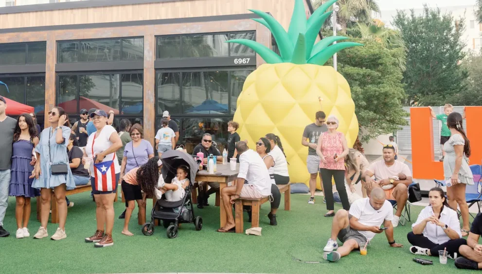 people outdoors sitting and standing around large inflatable pineapple, enjoying entertainment and food