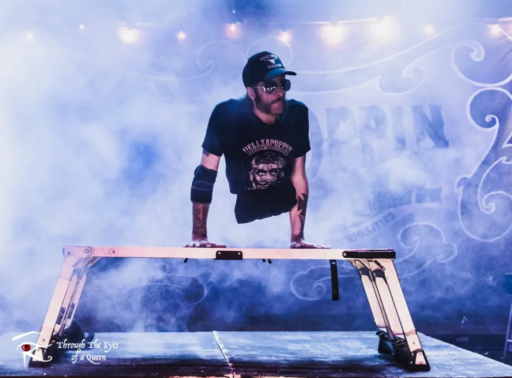Aaron Wollin, aka Short E. Dangerously, wearing a Hellzapoppin black Tshirt and black Texas hat, holds his torso up on his hands on a platform on stage with fog machine creating atmosphere around him.