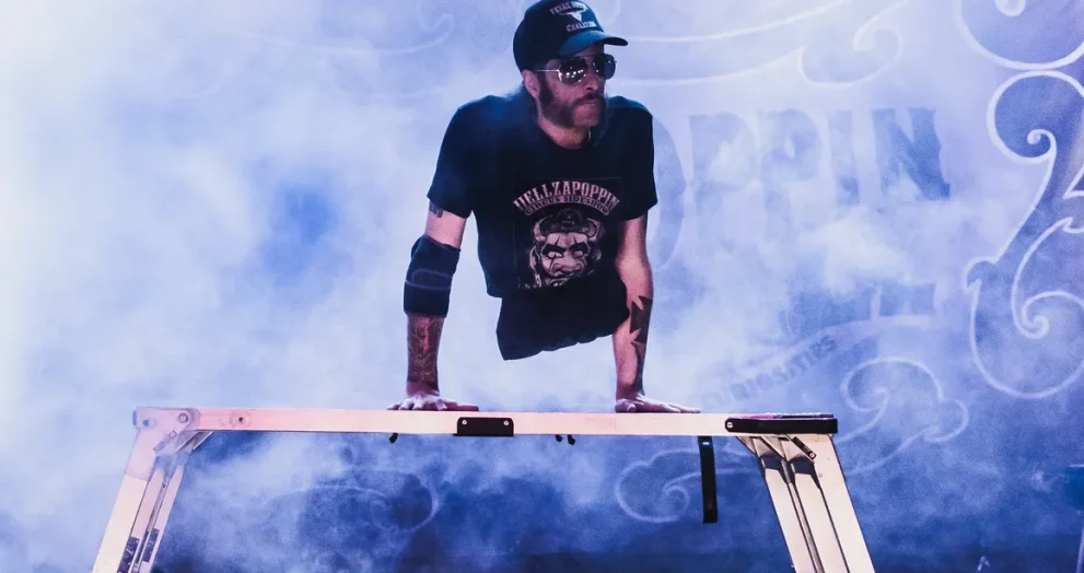 Aaron Wollin, aka Short E. Dangerously, wearing a Hellzapoppin black Tshirt and black Texas hat, holds his torso up on his hands on a platform on stage with fog machine creating atmosphere around him.