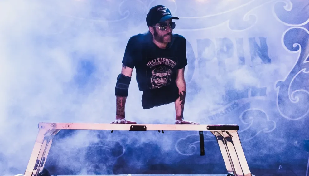 Aaron Wollin, aka Short E. Dangerously, wearing a Hellzapoppin black Tshirt and black Texas hat, holds his torso up on his hands on a platform on stage with fog machine creating atmosphere around him.