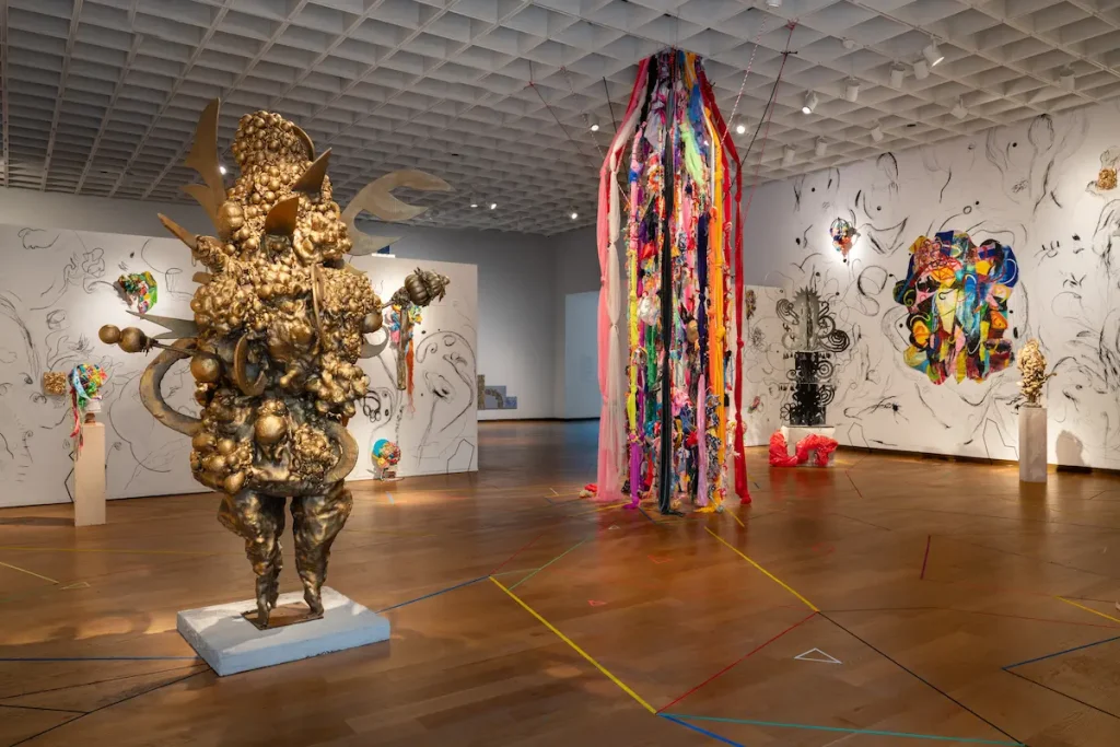 A gallery view of Charo Oquet's installation at the Orlando Museum of Art. The exhibition space features a large, textured gold sculpture on the left, a floor-to-ceiling installation of hanging colorful fabrics and ribbons in the center, and white walls covered in free-form black-and-white sketches interspersed with colorful mixed-media artworks. The wooden floor is decorated with intersecting geometric colored lines.