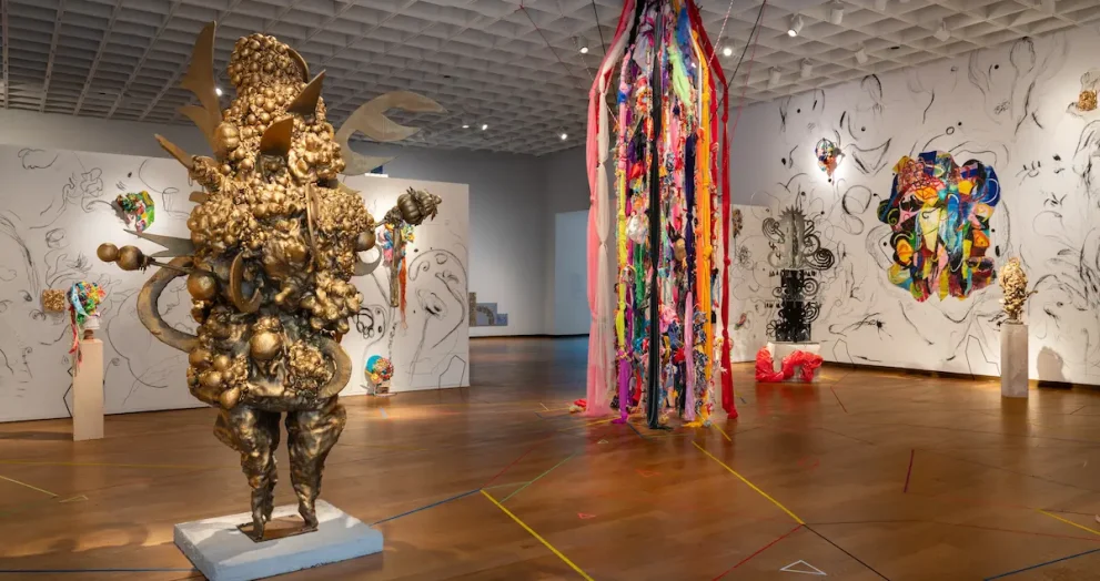 A gallery view of Charo Oquet's installation at the Orlando Museum of Art. The exhibition space features a large, textured gold sculpture on the left, a floor-to-ceiling installation of hanging colorful fabrics and ribbons in the center, and white walls covered in free-form black-and-white sketches interspersed with colorful mixed-media artworks. The wooden floor is decorated with intersecting geometric colored lines.