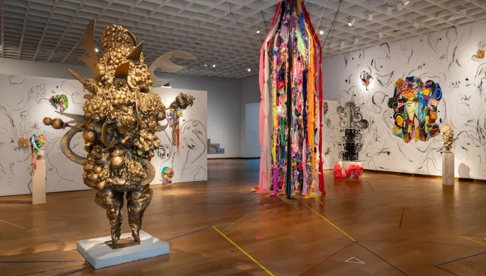 A gallery view of Charo Oquet's installation at the Orlando Museum of Art. The exhibition space features a large, textured gold sculpture on the left, a floor-to-ceiling installation of hanging colorful fabrics and ribbons in the center, and white walls covered in free-form black-and-white sketches interspersed with colorful mixed-media artworks. The wooden floor is decorated with intersecting geometric colored lines.