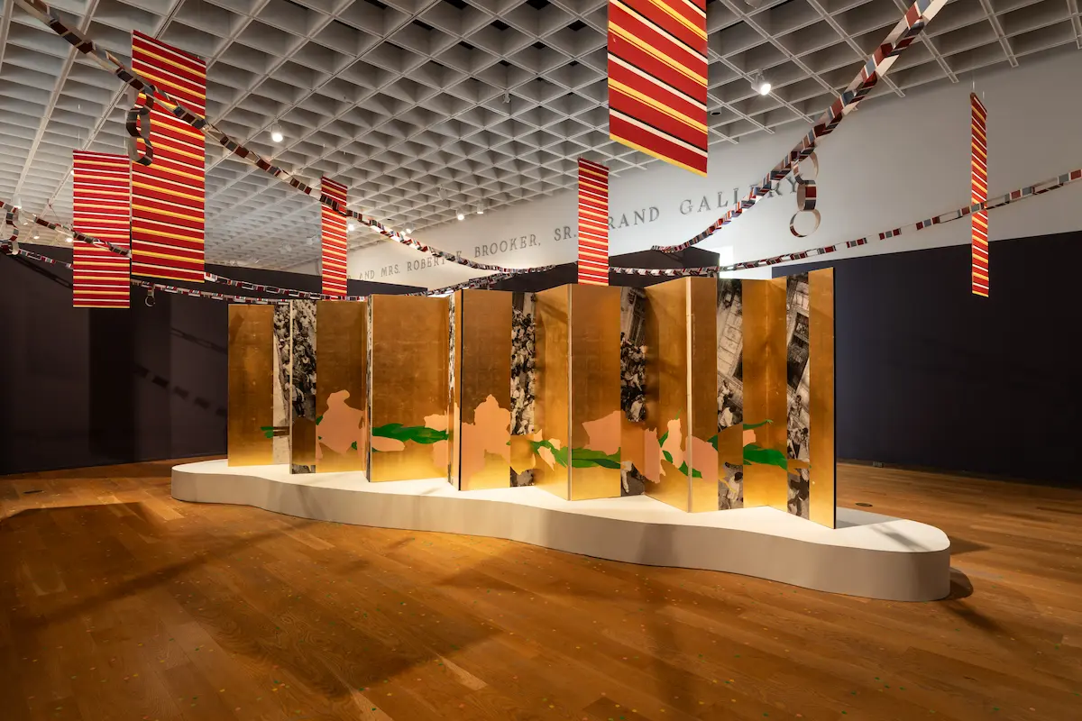 Inside Orlando Museum of Art, Francisco a large, accordion-style folding screen displayed on a low, curving white platform. The screen's panels alternate between reflective gold surfaces and black-and-white photographs of crowds. A continuous, abstract shape painted in pale pink and bright green stretches horizontally across the middle of the folding screen. Suspended from the gridded ceiling above are several rectangular banners with red, yellow, and dark horizontal stripes, along with long paper chains draped across the room.