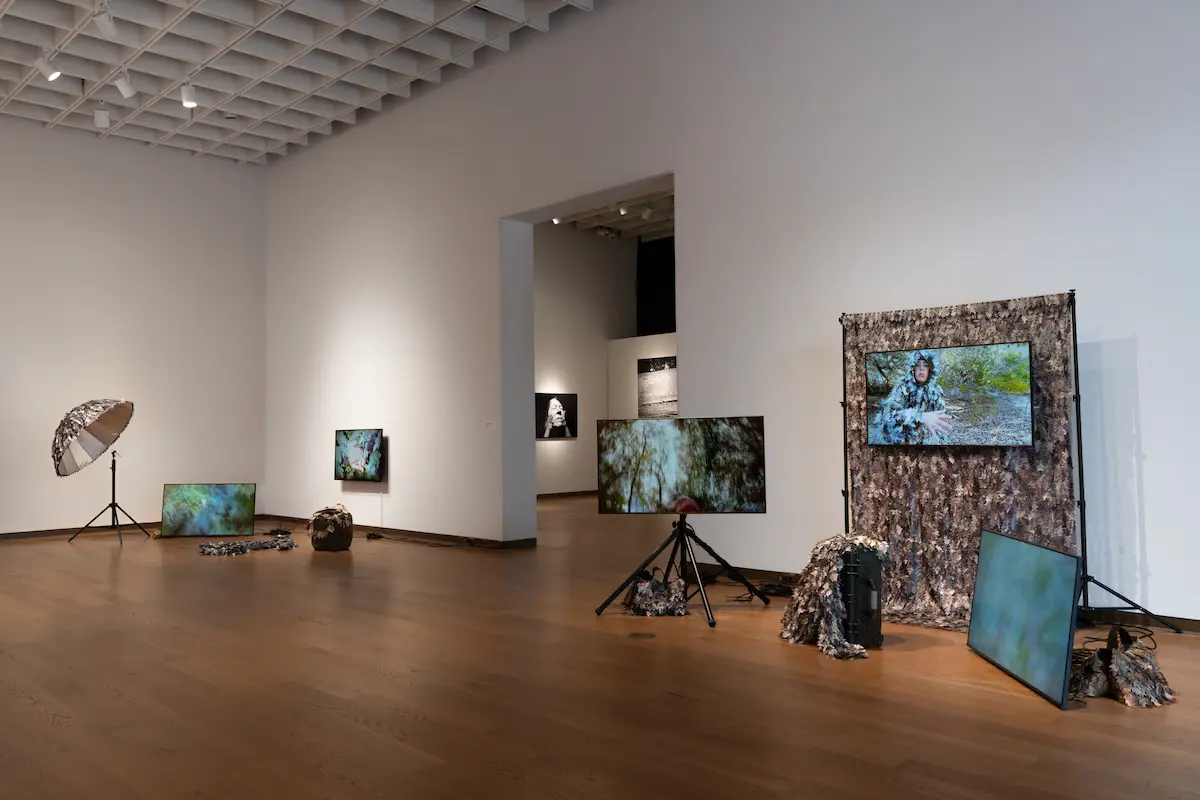 Orlando Museum of Art gallery with hardwood floors and white walls displaying a multimedia installation. Several flat-screen monitors show nature and camouflage-themed video art. The screens are positioned on tripods, resting on the floor, and mounted against a backdrop draped in camouflage fabric, accompanied by photography equipment. Another gallery room displaying black-and-white portraits is visible through an open doorway in the background.
