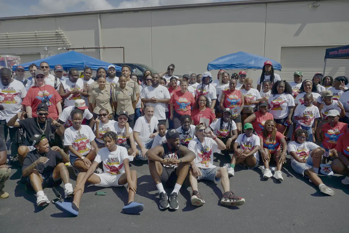 A wide, eye-level photo of a large, diverse crowd of dozens of children, teenagers, and adults gathering tightly together in a paved parking lot on a sunny day. Most of the volunteers are wearing matching custom t-shirts that feature colorful graphics and text reading "Back to School" and other matching uniforms. In the front row, several people are sitting cross-legged directly on the asphalt. The background features a plain, light-colored industrial concrete building wall with two blue pop-up canopy tents visible on either side of the crowd.