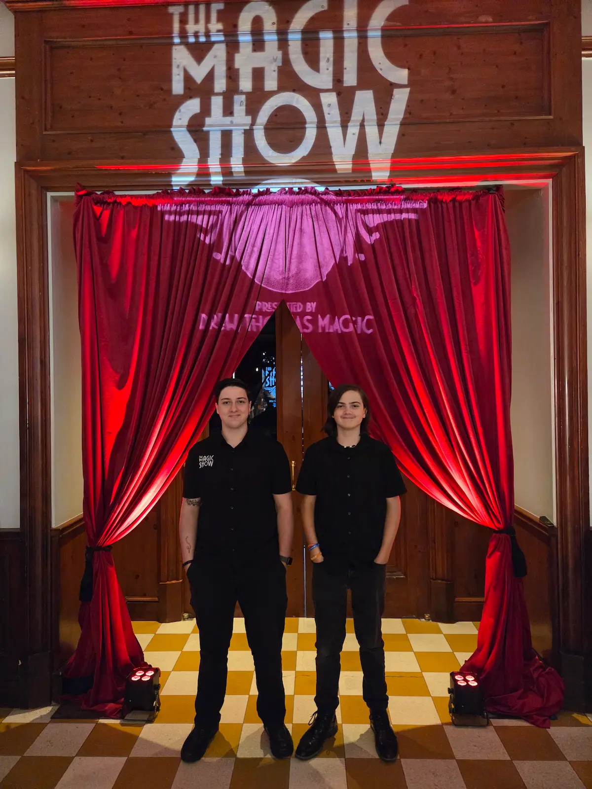 Sam and Milo Thomas are wearing black short-sleeved shirts and black pants and stand near the entrance to a venue which has red curtains pulled back and a spotlight above their heads. The floor has a yellow and orange checkerboard. The spotlight projects the words in white "The Magic Show" above the heads of the two young men.