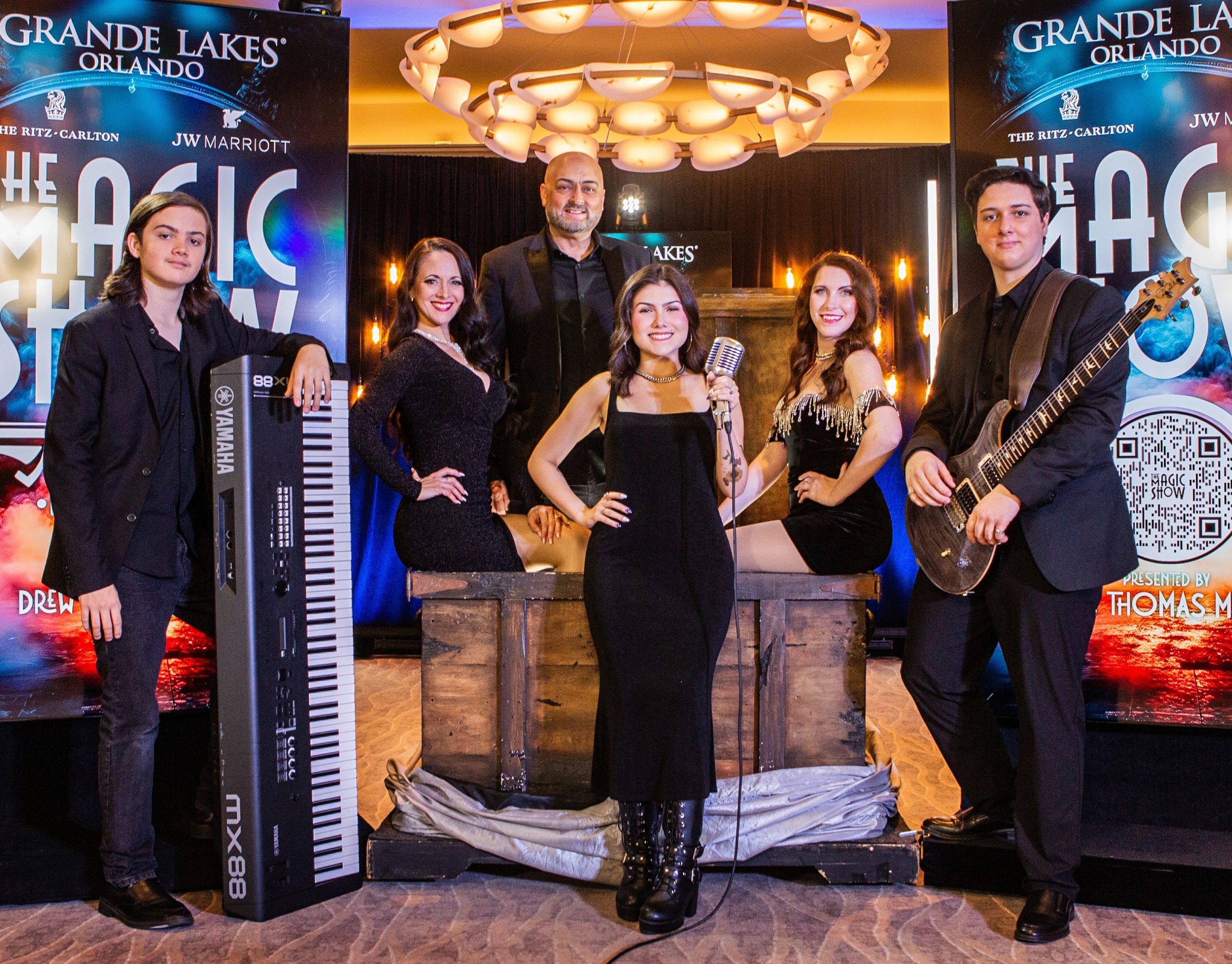 A group portrait of six performers—three men and three women—posing in formal black attire on a stage with musical instruments and a vintage microphone in front of a "The Magic Show" banner at Grande Lakes Orlando.