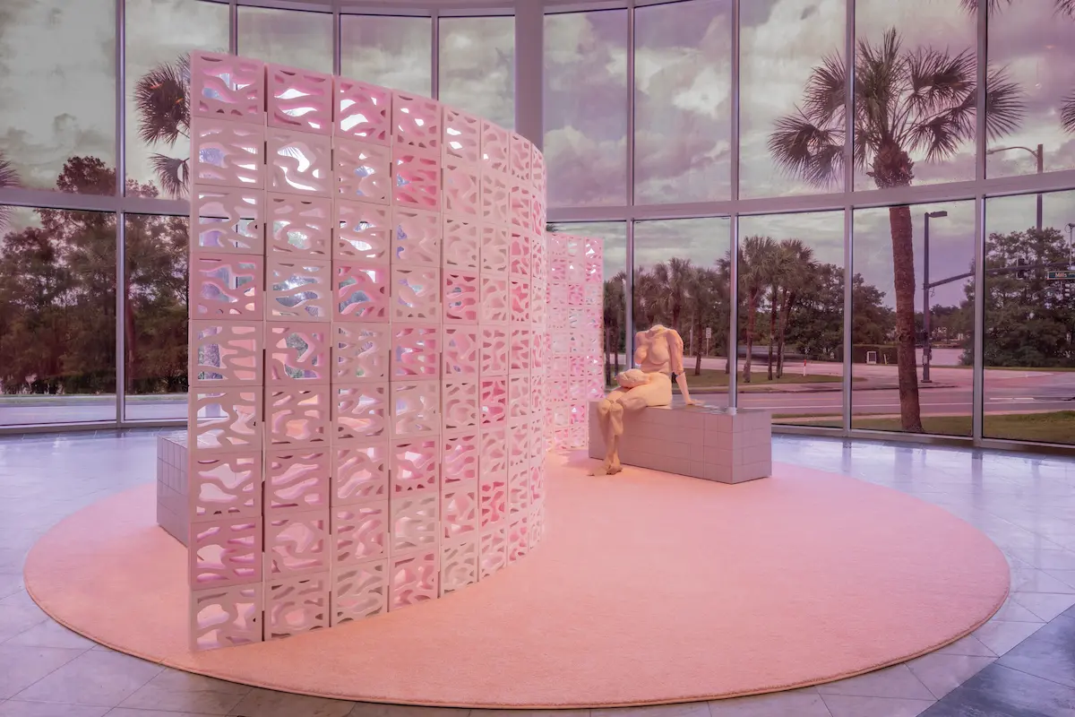 Art installation by We Are Nice'n Easy inside Orlando Museum of Art, with large floor-to-ceiling windows looking out onto a street with palm trees. A large, circular pink rug on the floor supports a curved wall made of decorative, pink breeze blocks. Behind the wall, a pale, headless sculpture of a seated female figure rests on a low, tiled bench, holding her detached head in her lap. The entire room is bathed in a soft, pink light.An obscured figure sits on the other side of the wall.