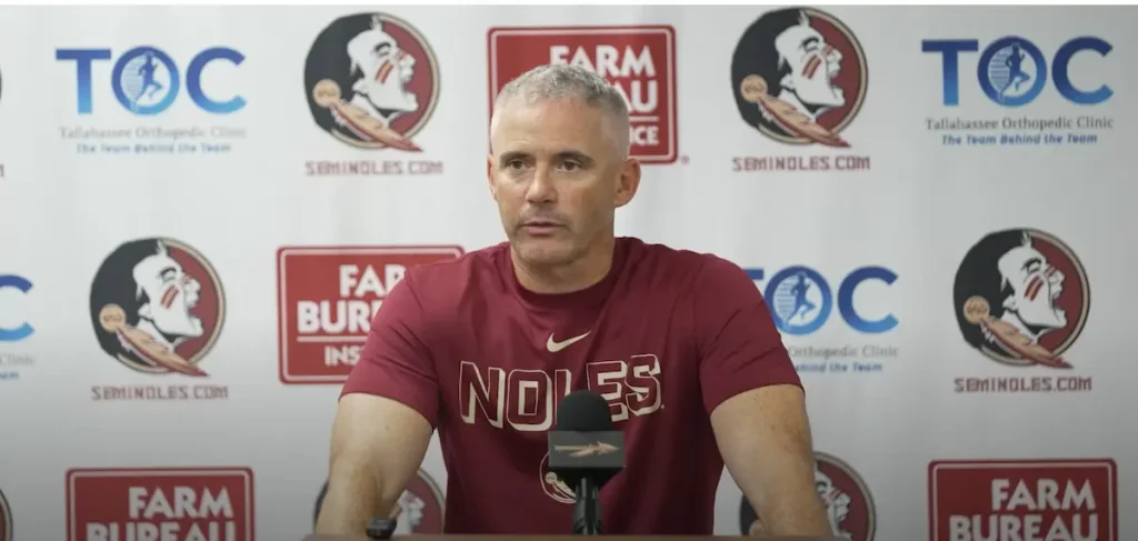 FSU Head Coach Mike Novell wears a garnet t-shirt that says "Noles" in capital letters and has a Nike swoosh at press conference before 2026 season opener. The background features the logos of sponsors including Seminoles.com, Tallahassee Orthopedic Clinic, and Farm Bureau Insurance