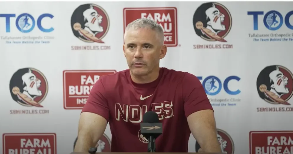 FSU Head Coach Mike Novell wears a garnet t-shirt that says "Noles" in capital letters and has a Nike swoosh at press conference before 2026 season opener. The background features the logos of sponsors including Seminoles.com, Tallahassee Orthopedic Clinic, and Farm Bureau Insurance