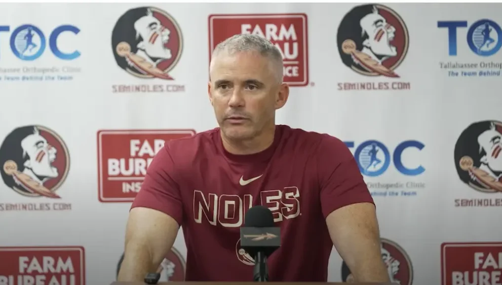 FSU Head Coach Mike Novell wears a garnet t-shirt that says "Noles" in capital letters and has a Nike swoosh at press conference before 2026 season opener. The background features the logos of sponsors including Seminoles.com, Tallahassee Orthopedic Clinic, and Farm Bureau Insurance