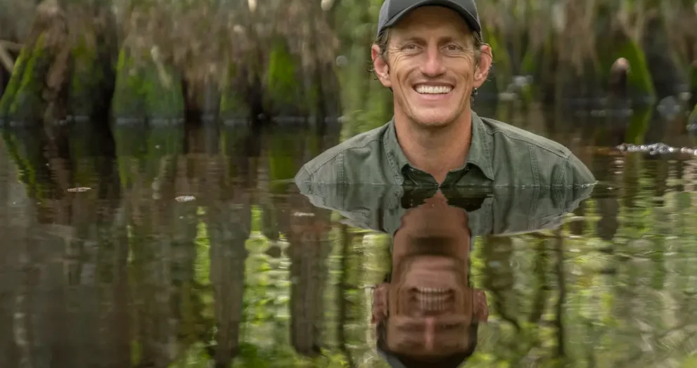 Nat Geo Explorer and photographer Carlton Ward, Jr. is submerged in a swamp up to his shoulders. He wears a green hat and green shirt and smiles at the camera. His reflection can be seen in the water. Cypress knees rise up from the water behind him.