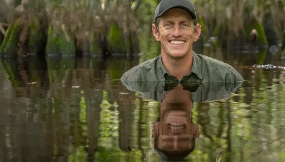 Nat Geo Explorer and photographer Carlton Ward, Jr. is submerged in a swamp up to his shoulders. He wears a green hat and green shirt and smiles at the camera. His reflection can be seen in the water. Cypress knees rise up from the water behind him.