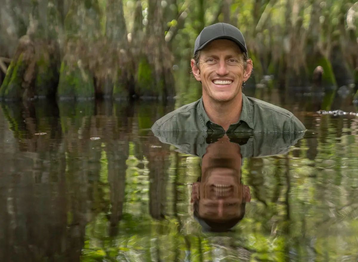 Nat Geo Explorer and photographer Carlton Ward, Jr. is submerged in a swamp up to his shoulders. He wears a green hat and green shirt and smiles at the camera. His reflection can be seen in the water. Cypress knees rise up from the water behind him.