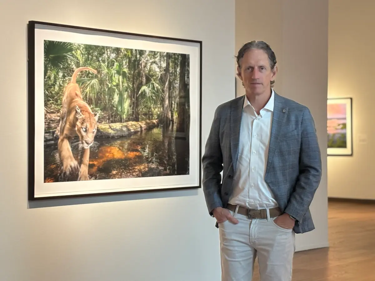 Carlton Ward, Jr. stands in front of a framed large print of his 2017 photo titled Log Jump, which features a Florida panther in a swamp of the Florida Panther Wildlife Refuge. Ward wears a blue jacket and white shirt, with tan jeans and a brown belt.