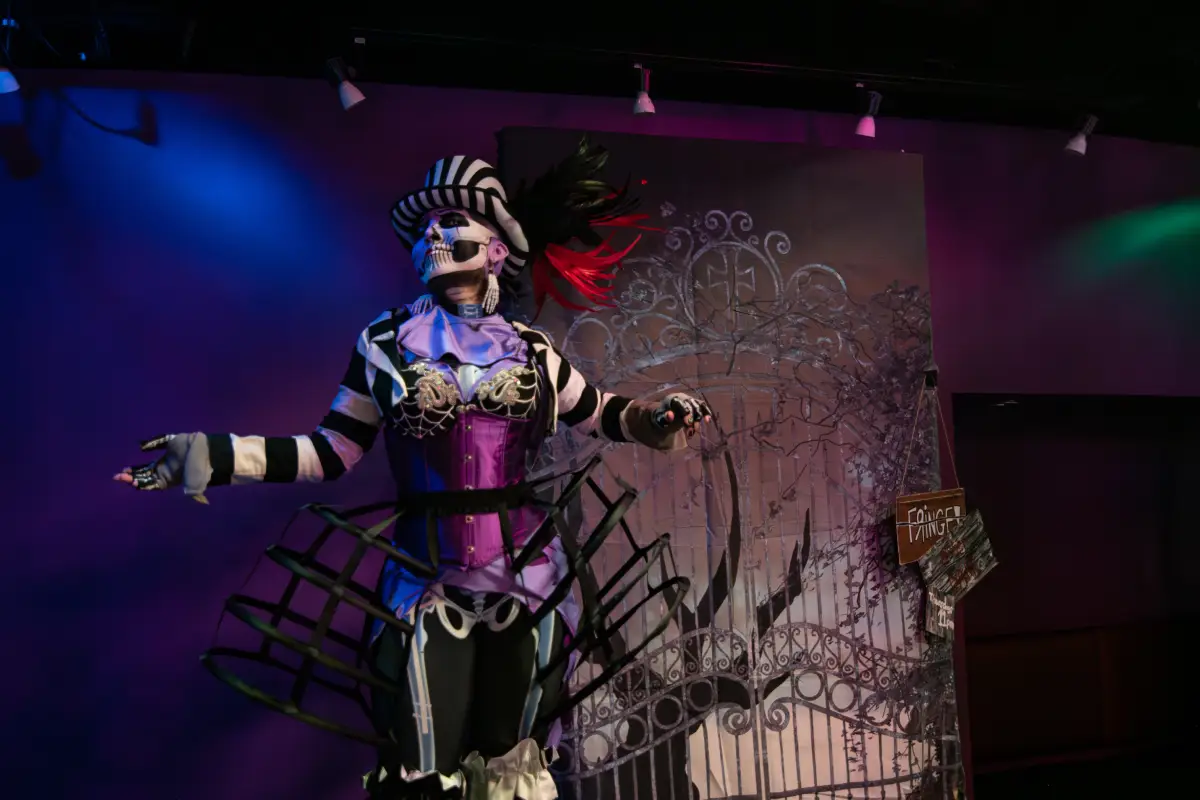 Performer in a theatrical skeleton costume, purple corset, and hoop skirt cage on a stage with purple lighting and a cemetery gate backdrop.