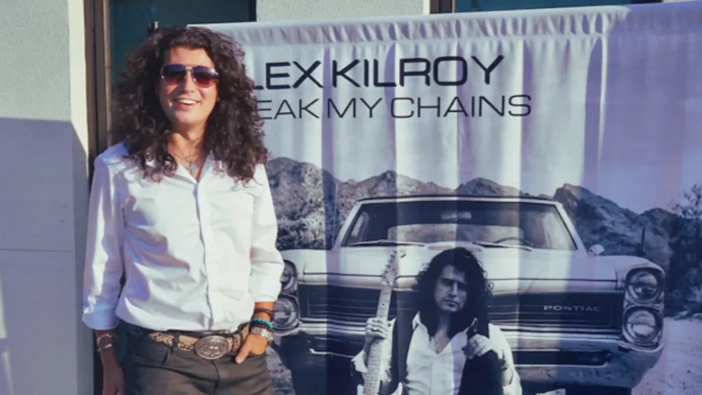 Guitarist Alex Kilroy standing in front of his debut album promotional poster