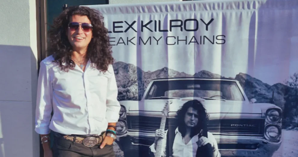 Guitarist Alex Kilroy standing in front of his debut album promotional poster