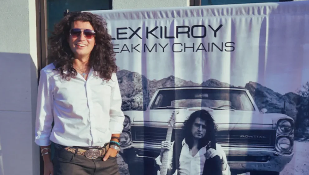 Guitarist Alex Kilroy standing in front of his debut album promotional poster